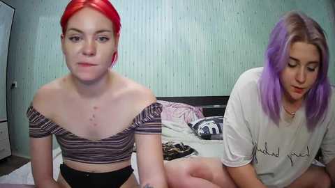 Media: Video of two young women with pale skin; one with red hair, wearing a striped off-shoulder crop top, and the other with purple hair, wearing a white T-shirt. Both sitting on a bed in a room with light blue walls and a white dresser.