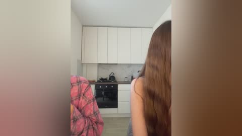 Media: Video of a woman with long brown hair sitting in a modern kitchen, wearing a pink plaid shirt, facing away. White cabinets, stainless steel appliances, and a grey backsplash are visible.