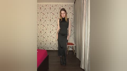 Video of a pregnant woman in a black sleeveless dress, standing in a minimalistic room with white floral wallpaper and a white chair.