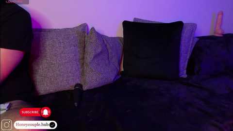 A video showing a woman's legs and black dress on a dark bed, with gray and black pillows, under purple lighting.