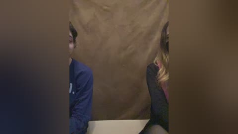A video of two individuals sitting back-to-back in a dimly lit room, obscured by a brown curtain. The man on the left wears a blue shirt, while the woman on the right has blonde hair and a dark top.