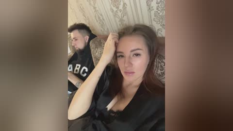 Media: Video of a young Caucasian woman with brown hair and a man with a beard and short hair, both wearing black robes, seated on a bed with beige, patterned wallpaper.