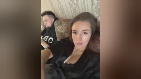Media: Video of a woman with fair skin, brown hair, wearing a black robe, sitting on a brown couch, next to a man in a black shirt with a white logo.