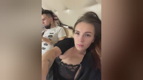 Media: Video of a man and woman in a car. Man with tattoos, short hair, and beard, wearing a white t-shirt. Woman with long brown hair, light skin, wearing black lace bra, and black jacket, looking at the camera.