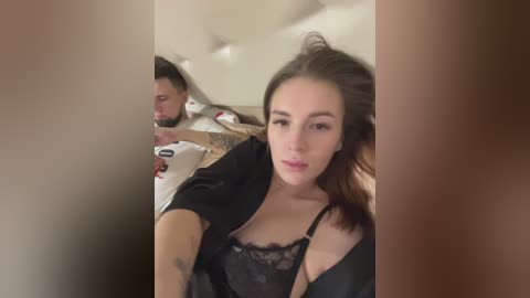 Media: Video of a young woman with light skin, brown hair, and a black lace bra, sitting on a bed next to a man in a white shirt. The background is a beige upholstered headboard.