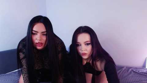 Media: Video of two women with long black hair, dressed in black lace outfits, posing provocatively on a bed in a dimly lit room.