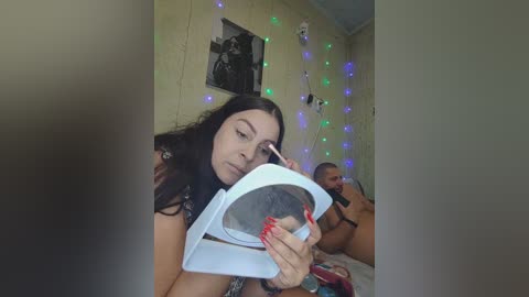 A candid video shows a young woman with long black hair, applying makeup in a bedroom adorned with string lights and a framed picture.