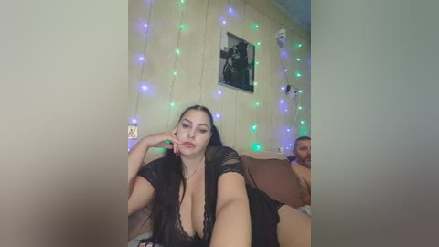 Video of a plus-sized woman with fair skin, dark hair, and red lipstick, wearing a black lace top, sitting on a brown couch. Behind her, a beige wall with string lights and a framed photo.
