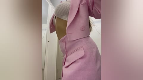Video of a person in a pink coat, adjusting a white knit hat, standing in a white-walled room with a door visible in the background.
