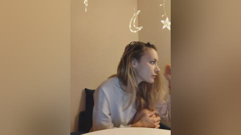 Media: A video of a young woman with light skin and long blonde hair, wearing a light-colored sweater, seated at a table, gazing into the distance, with moon and star decorations hanging in the background.