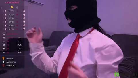 Video of a person wearing a black mask, white shirt, and red tie, gesturing, in a dimly lit room with a gray couch, a screen displaying \"LOVEWAVES\" and chat options.