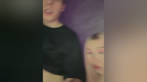 Media: Video of a blurry scene featuring a person with light skin wearing a black top and another person with pink makeup, possibly a woman, partially visible. Background is indistinct and dark.