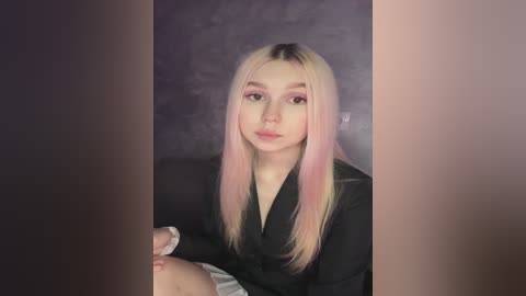 Media: Video of a young woman with long, pastel pink and blonde hair, fair skin, and subtle makeup, seated on a dark couch. She wears a black blazer.