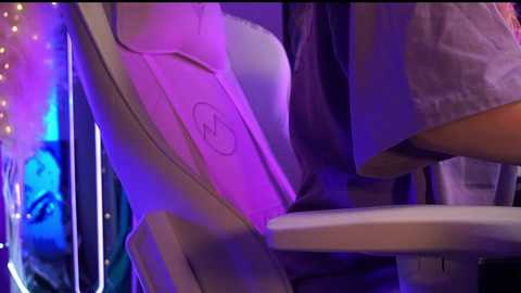 Media: A video of a person sitting in a modern, purple-lit chair with a white cushion, wearing a light gray t-shirt, with the background showing a blurred, fairy-lit forest scene.