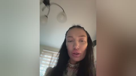 Video of a young woman with long black hair, fair skin, and full lips, standing in a room with a chandelier, white walls, and a window with closed blinds.