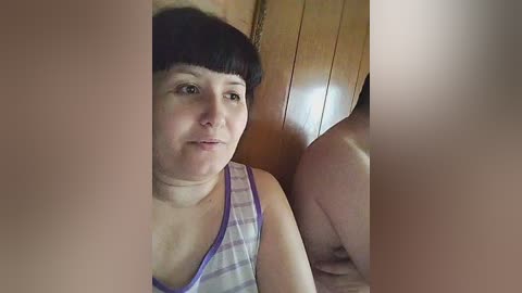 Video of a fair-skinned woman with short black hair and bangs, wearing a plaid tank top, sitting in a wooden room with another person's blurred arm visible.
