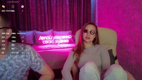 Video of a young woman with long brown hair, wearing sunglasses, sitting on a cream-colored couch with a neon light behind her. A man in a blue shirt is partially visible on the left.
