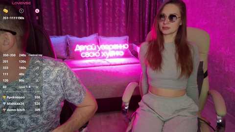 Video of a young woman with long brown hair and large sunglasses, wearing a grey crop top and high-waisted pants, sitting on a beige chair in a dimly lit room with pink neon lights and a couch behind her.