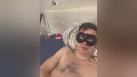 Media: Video of a man with short dark hair, wearing a black mask, lying on a bed with white pillows and a polka-dotted blanket, in a dimly lit room.
