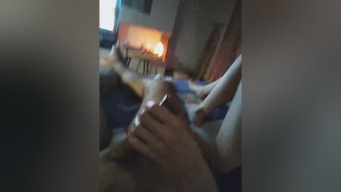 A blurry video captures a dimly lit, intimate indoor scene featuring a man's hand stroking a woman's exposed, large breasts. The background shows a wooden dresser and a lit fireplace, creating a warm ambiance.