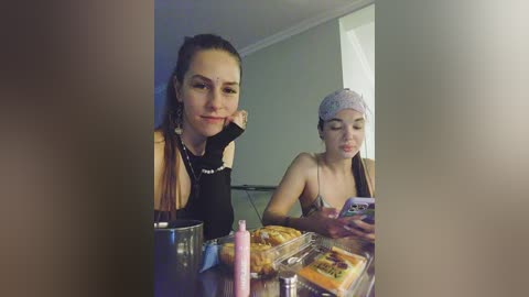 Video of two young women seated at a table, one with dark hair, black top, and a pink bottle, the other with a headscarf, wearing a tank top, eating a sandwich.