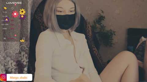 A video of a fair-skinned woman with straight, shoulder-length gray hair, wearing a white blazer, black face mask, and seated in a dark, patterned armchair.