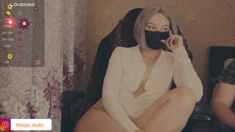 A video of a young woman with light skin, long straight hair, and a black face mask, wearing a sheer white blouse and revealing her cleavage, sitting on a black chair in a cozy room with floral wallpaper.