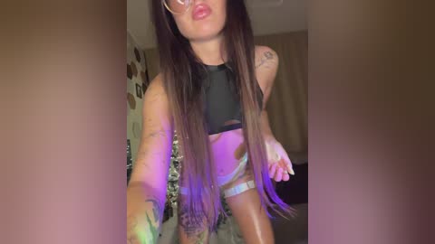 Video of a young woman with long dark hair, wearing glasses, black crop top, and white panties, posing in a dimly lit room with purple and green lights, tattoos on her arms.