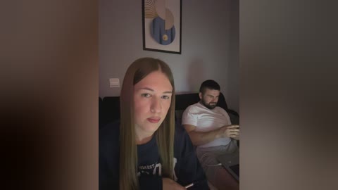 Media: Video: A young woman with long straight hair and a white shirt sits on a dark couch, looking slightly to the left. Behind her, a man with a beard sits in a white shirt, focused on a phone.