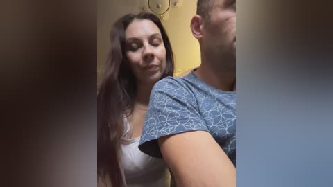 Media: Video of a smiling woman with long brown hair and a white top, hugging a man with short brown hair, wearing a blue patterned shirt, in a dimly lit room.