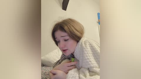 Video of a young woman with fair skin and light brown hair, wearing a white, textured robe, sitting on a bed with a floral-patterned pillow, appearing to be sick with a green face mask on.