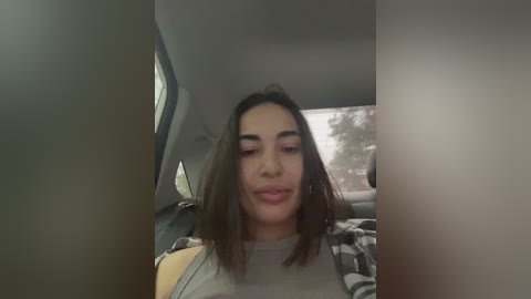 Media: Video of a young woman with medium brown skin and shoulder-length brown hair, wearing a gray top, seated in a car.