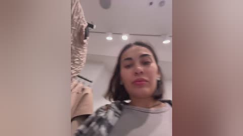 Media: Video of a young woman with medium skin tone, dark hair, and full lips, wearing a gray shirt, standing in a minimalist room with beige walls and recessed lighting.