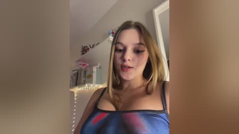 Media: A video of a young, light-skinned woman with long blonde hair, wearing a low-cut blue top, standing indoors near a bookshelf with decorative items and a string of lights.