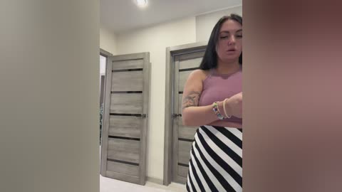 Video of a curvy woman with long black hair, wearing a pink top and black-and-white striped skirt, standing in a modern hallway with grey doors.