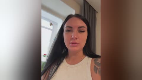 A video of a young woman with long black hair, wearing a white tank top, standing indoors near a window with a blurred view of greenery outside.