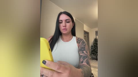 A video of a tattooed woman with long black hair, wearing a white tank top, taking a selfie in a beige hallway.