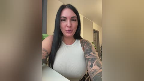 Video of a tattooed, light-skinned woman with long black hair in a white ribbed tank top, indoors, blurred background.
