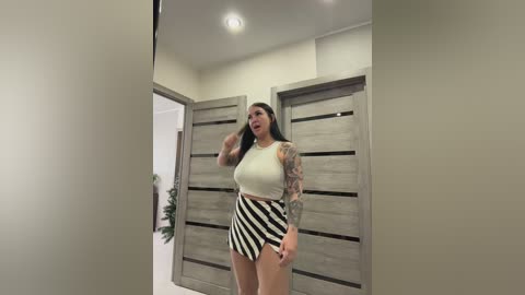 A video of a tattooed woman with long dark hair, wearing a white crop top and black and white striped skirt, standing in a modern, beige-walled hallway.