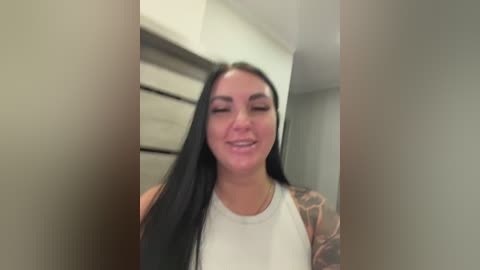 Video of a smiling young woman with long black hair, wearing a white sleeveless top, standing in a modern, minimalist hallway with beige walls and a wooden staircase.