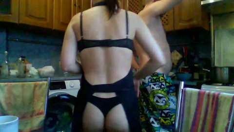 Video of a woman in black lingerie, back view, standing in a kitchen. She has fair skin, medium build, and is wearing a black bra and thong. Background features wooden cabinets, a striped towel, and kitchen items.