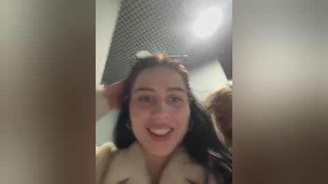 Media: Video of a smiling young woman with long brown hair, wearing a beige fur-trimmed coat, captured in a dimly lit room with a checkered patterned ceiling.
