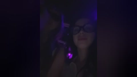 Media: A dimly lit video of a woman with glasses, wearing a black top, holding a purple-tinted flashlight, her face partially obscured.