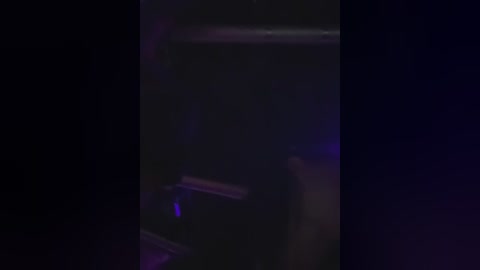 Media: A dimly lit, blurry video of a person standing in front of a dark, possibly metallic, object with indistinct features. The lighting casts a purple hue, creating a mysterious and slightly eerie atmosphere.
