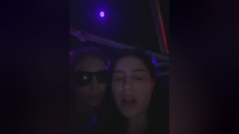 Media: A dimly lit video captures two women in a car at night, one with sunglasses, the other with closed eyes, both with pale skin and dark hair. The car's interior is dark, with purple light from the dashboard.