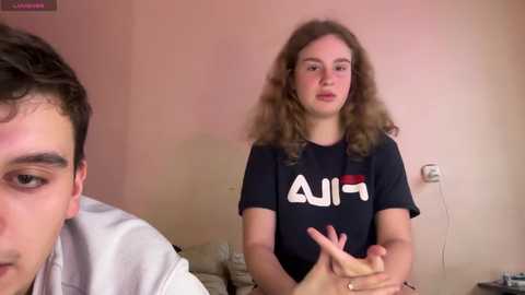 Media: Video of a young Caucasian woman with curly hair, wearing a black shirt with white text, and a man in the foreground. Both seated in a living room with beige walls.