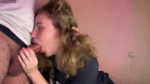 Media: Video of a young Caucasian woman with curly brown hair performing oral sex on a man with a large, erect penis, both in a room with beige walls and a light switch.