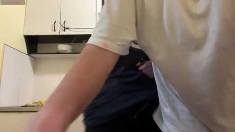 Media: Video of a person in a white shirt and dark pants bending over in a kitchen with white cabinets, a yellow dish, and a black handle.
