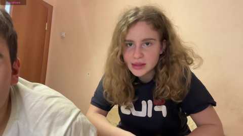 Media: Video of a young woman with curly blonde hair and fair skin, wearing a black shirt with \"SUI\" text, looking concerned while a man in a white shirt is in the background.