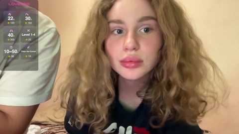 Media: Video of a young, pale-skinned girl with long, wavy blonde hair, wearing a black shirt, looking directly at the camera. Background shows a beige wall with a digital clock and thermometer display.